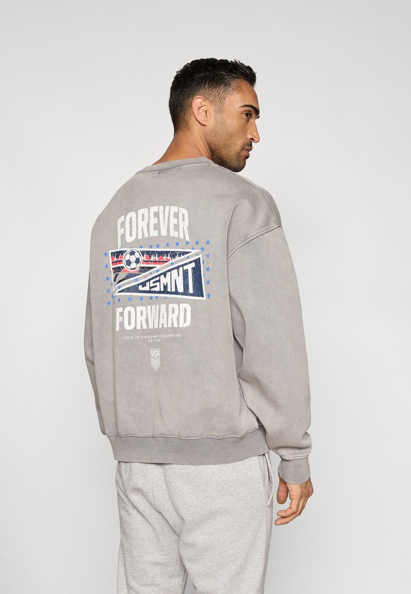 Γκρι φούτερ με άνετη εφαρμογή, διαθέτει γραφικό σχέδιο στην πλάτη με τη φράση «FOREVER FORWARD» και λογότυπο USMNT σε μπλε και κόκκινες αποχρώσεις.
