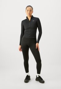 Musta vetoketjulla varustettu urheilu paita pitkällä hihalla, yhdistettynä mustiin leggings-housuihin. Näyttää sileältä kankaalta ja istuvalta muotoilulta. Nike-logo rintamalla.