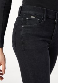 Dark denim jeans with a fitted design, featuring multiple pockets and a G-Star label. The fabric has a smooth texture and subtle sheen.