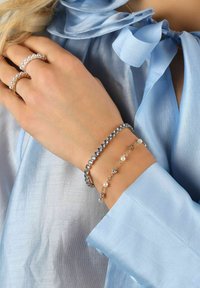 Silver- och guldarmband med blå kristaller och pärlor, som bärs på en hand. Ljusblå satinblus synlig i bakgrunden.