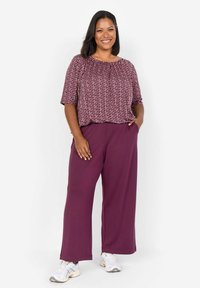 Pantaloni larghi viola abbinati a una maglietta a maniche corte color bordeaux con motivi geometrici. Il modello indossa scarpe da ginnastica bianche.