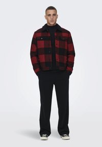Jachetă flanelată cu carouri roșii și negre, cu guler negru, purtată cu un tricou negru cu mânecă lungă și pantaloni negri, completată cu încălțări casual.