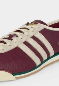 Μπορντό αθλητικό παπούτσι Adidas με λευκές δερμάτινες ρίγες, λευκά κορδόνια, υφή στη σόλα και χρυσό λογότυπο στη γλώσσα και την πλευρά.