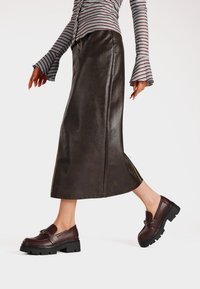 Gonna lunga in finta pelle marrone abbinata a mocassini platform bordeaux con una fibbia decorativa e suola testurizzata. Maglietta a righe con maniche svasate.