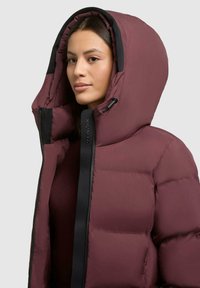 Giacca a vento bordeaux con grande cappuccio, chiusura a zip e dettagli neri. Presenta una texture trapuntata e un marchio sulla parte anteriore.