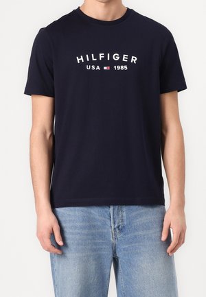 Hombre con camiseta azul marino con “HILFIGER USA 1985” y logo de bandera roja, blanca y azul, combinado con vaqueros azul claro.