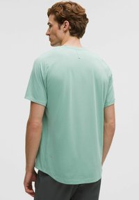 Tricou athletic de culoare verde deschis, cu uscare rapidă, cu mâneci scurte, decolteu rotund și un logo mic aproape de margine, cu textură de țesătură netedă.