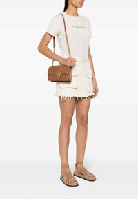 Sac en bandoulière en cuir marron matelassé avec un accent en forme d'anneau doré. Modèle portant un T-shirt blanc, une jupe en denim et des sandales marron.
