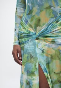 Nahaufnahme einer Person, die ein langärmeliges, grün-blaues Blumenkleid mit einem verdrehten Knoten an der Taille und einem hohen Schlitz im Rock trägt.