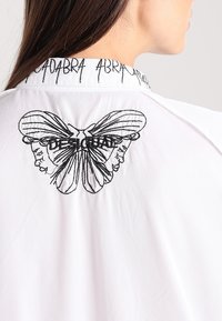 Camicia bianca con ricami neri di una farfalla e volti sul retro, con il testo "DESIGUAL" integrato nel design.