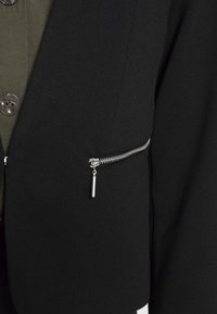 Veste noire en tissu texturé, design élégant, poche zippée avec matériel argenté, portée sur une chemise verte avec des boutons ronds.