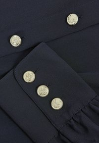Black fabric with three gold-tone buttons featuring embossed designs. Smooth texture with detailed stitching and a gathered accent.