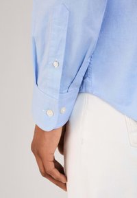 Lichtblauw blouse met lange mouwen en knoopmanchetten. De manchetten zijn voorzien van twee met stof bedekte knopen. Zachte textuur, getailleerde pasvorm, gedetailleerd gestikt.