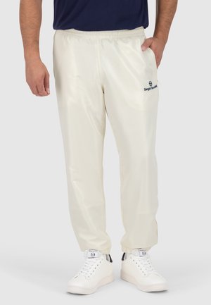 Homme portant un jogging écru à taille élastique et des baskets blanches avec des languettes noires au talon sur un fond clair uni.