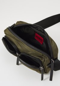 Olivgrüne Nylon-Crossbody-Tasche mit zwei Reißverschlussfächern, schwarzen Reißverschlüssen und Ziehgriffen, mit einem roten Logo-Tag innen.
