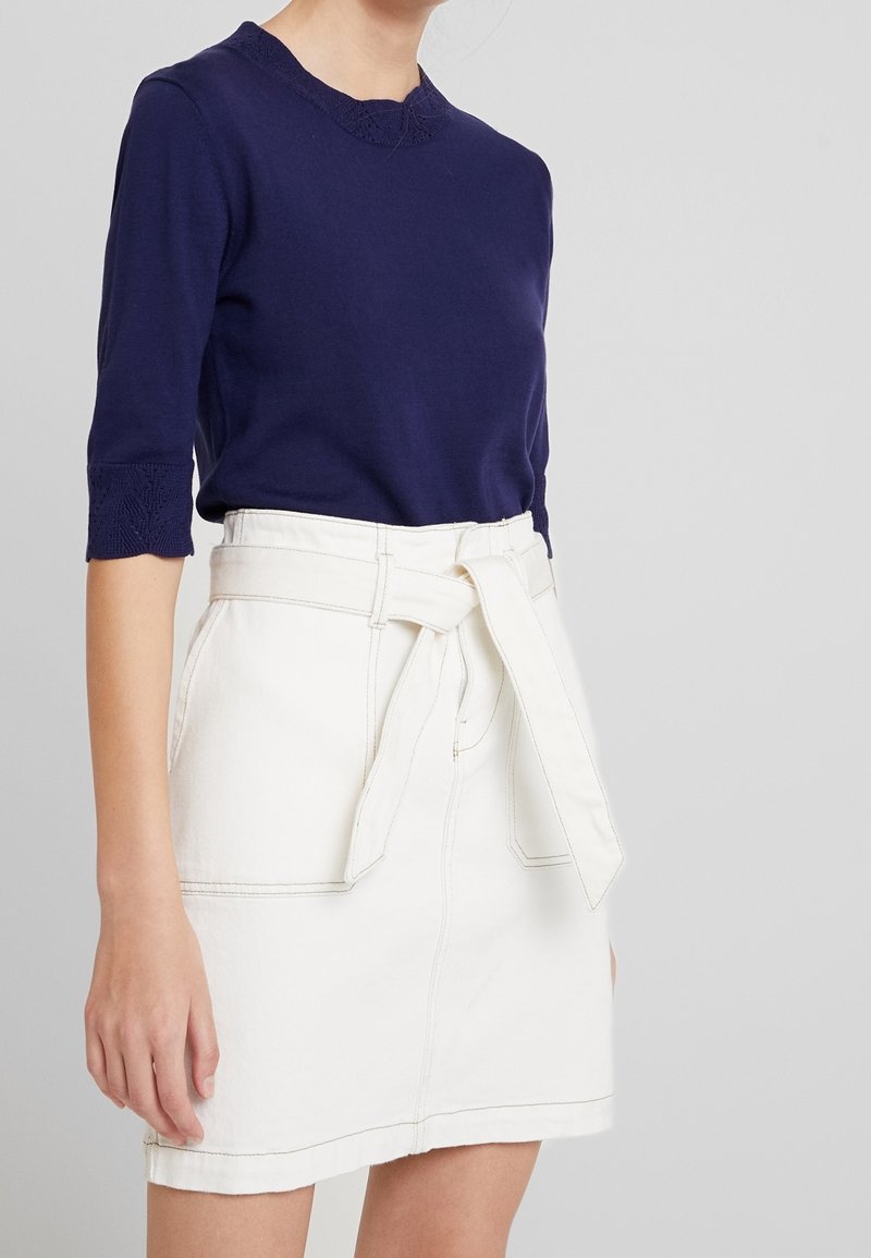 Námořnicky modrý pletený top s 3/4 rukávy a krajkovým detailem na výstřihu, kombinovaný s bílou džínovou sukní se zavazovacím pásem a kapsami.