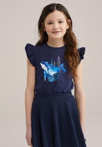 Námořnická modrá tričko s volánovými rukávy, se grafikou modré velryby a mořskými prvky. V páru s texturovanou námořnickou sukní.