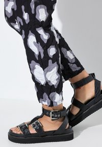 Schwarze Plateau-Sandalen mit strukturiertem Krokodilmuster, verstellbaren Riemen, Metall-schnallen und einer dicken Sohle. Kombiniert mit gemusterten Hosen.