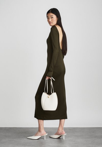 Abito lungo verde oliva con maniche lunghe e schiena scoperta, abbinato a una borsa bianca e mule di colore chiaro, su uno sfondo neutro.