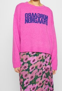 Pull en tricot rose au design court avec des manches larges, présentant un texte bleu audacieux "NORGAAARD" tricoté sur le devant.