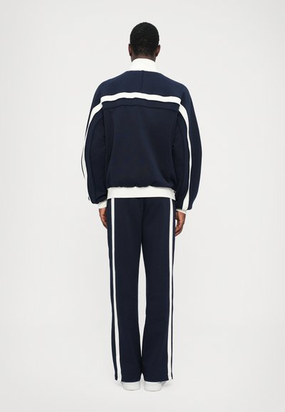 Set de hanorac și pantaloni bleumarin cu dungi albe pe lateral, caracterizat printr-o croială lejeră și manșete ribbate, fabricat dintr-un material moale și texturat.