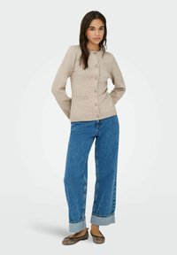 Cardigan en tricot beige avec un col rond et des boutons, associé à un jean bleu clair à jambes larges et des chaussures plates marron.