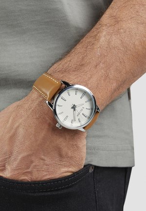 Silberne Uhr mit weißem Zifferblatt und silbernen Markierungen, ausgestattet mit einem braunen Lederarmband mit kontrastierender Naht, getragen am Handgelenk.