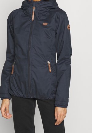 Person wearing a navy blue hooded jacket with brown zipper pulls and patches, standing against a plain light background.