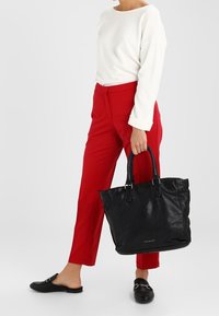 Sac fourre-tout en cuir noir avec anses doubles, finition texturée et accent de logo argenté, assorti à un pantalon rouge et un haut à manches longues blanc.