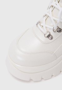 Botine albe chunky cu un finisaj lucios, având eyeleturi metalice, șireturi albe și o talpă groasă, texturată. Partea superioară este fină, având detalii de cusătură.