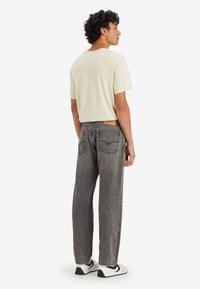 Camiseta de color beige claro para hombre combinada con jeans gris de tiro alto y corte holgado, que presentan una etiqueta de cuero y costuras en contraste, junto con zapatillas deportivas.