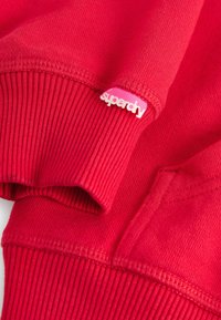 Červená mikina s ribovaným lemem a manžetami, na manžetě malá růžová nálepka s nápisem "superdry". Měkká textura a standardní střih.