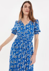 Vestido floral azul con mangas cortas abullonadas, cintura fruncida y un profundo escote en V. Presenta patrones de flores blancas y negras en una tela suave.