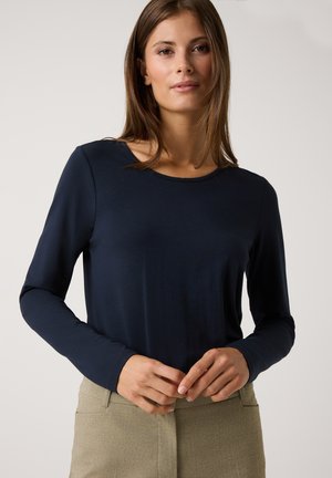 Marineblaues Langarm-Oberteil aus glattem Stoff mit rundem Ausschnitt. Das Modell trägt hellbeige Hosen und hat die Hände in der Taille positioniert.