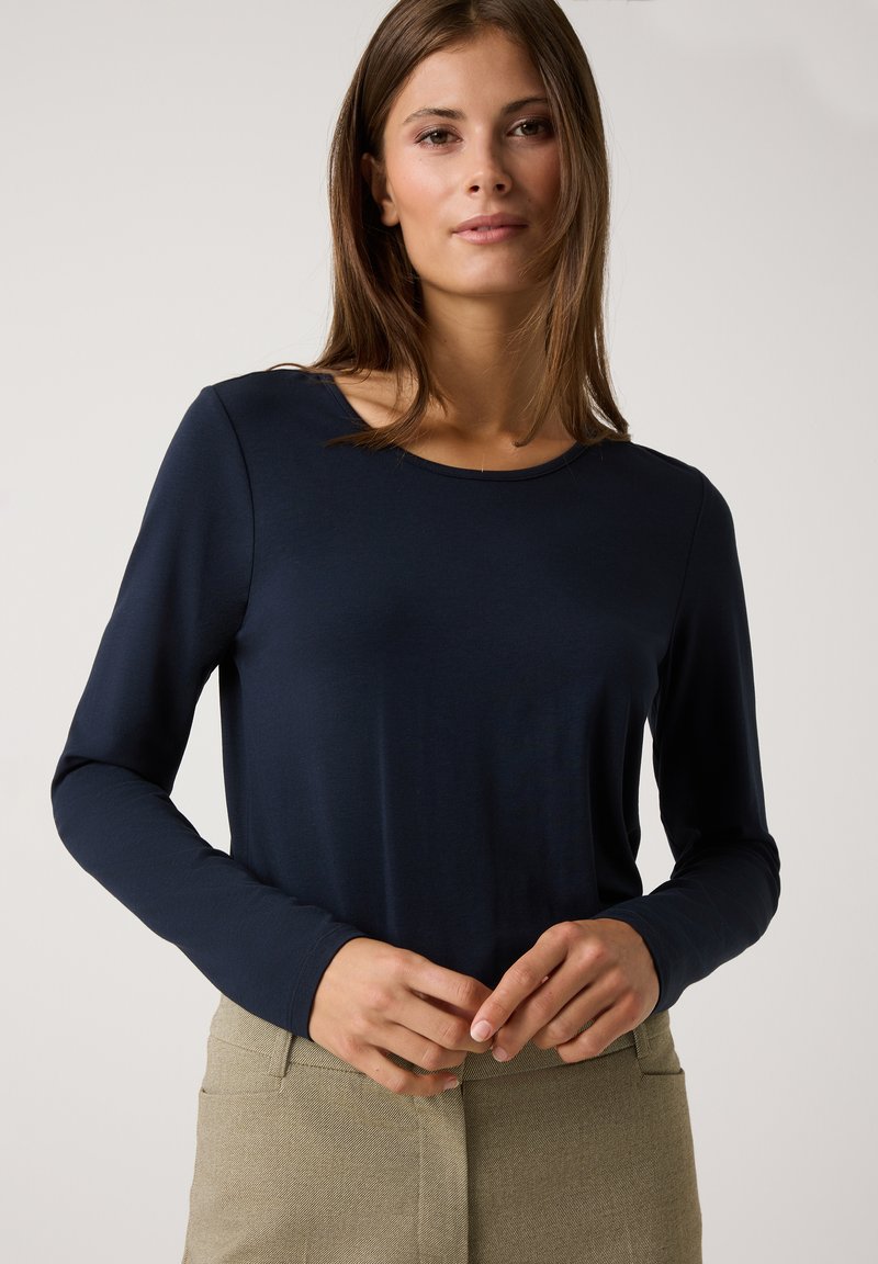 Marineblauwe top met lange mouwen van gladde stof en een ronde halslijn. Het model draagt een lichtbeige broek en heeft de handen op de heupen geplaatst.