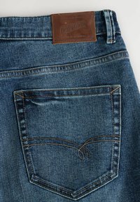 Tmavomodré džínsy z denimu s koženou nášivkou na zadnej strane a kontrastným prešívaním na vrecku. Majú tradičný dizajn s piatimi vreckami.