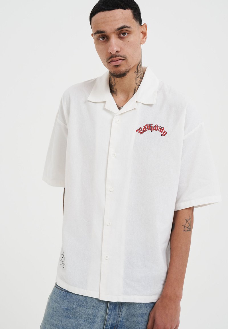 Korte mouwen witte blouse met een knoopsluiting aan de voorkant, zachte textuur en rode borduurtekst op de linkerborst. Casual pasvorm met zichtbare stiksels.