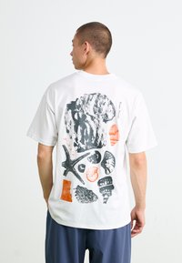 Man wearing white t-shirt with black and orange fossil and seashell illustrations on the back, standing against a plain white background.
