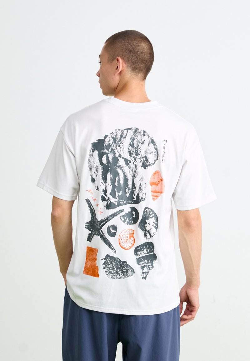 Man wearing white t-shirt with black and orange fossil and seashell illustrations on the back, standing against a plain white background.