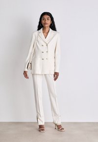 Beige double-breasted blazer with large pockets, paired with tailored pants. The outfit features gold-tone buttons and a smooth texture.