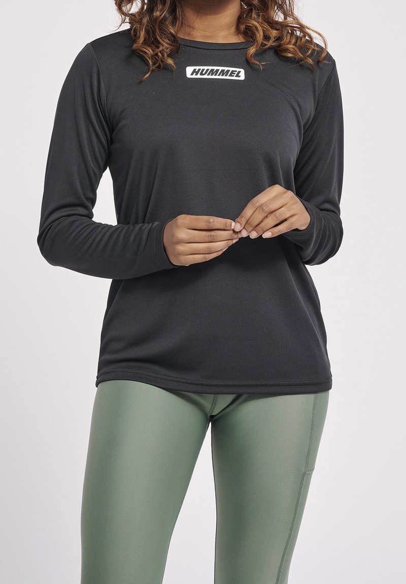 Schwarz langärmliges Sportshirt mit Rundhalsausschnitt und einem weißen "HUMMEL"-Logo auf der Brust, kombiniert mit enganliegenden grünen Leggings.