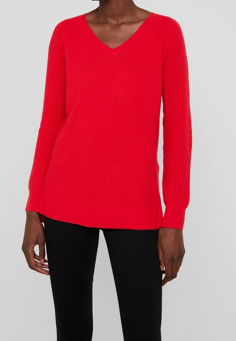 Personne portant un pull à manches longues col en V rouge vif et un pantalon noir sur un fond gris uni.