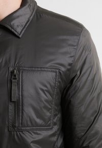 Chaqueta acolchada negra con bolsillo en el pecho con cremallera, cuello con solapa y textura de tela suave y brillante.