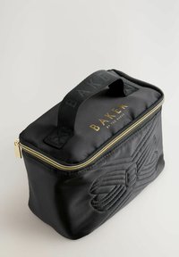 Borsa cosmetica nera trapuntata con zip dorata, caratterizzata da un design a fiocco embossato su un lato e un manico in tessuto nero. Logo "BAKER" visibile.