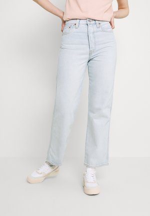 Personne portant un jean droit taille haute bleu clair, des baskets beige et blanches, et un haut rose clair rentré, debout les mains sur les hanches.