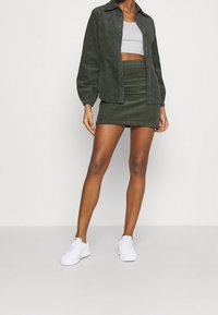 Camisa de pana verde, top gris corto, falda mini de canalé verde a juego y zapatillas blancas. El conjunto presenta un diseño de camisa con botones.