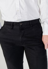 Černé bavlněné kalhoty s úzkým střihem, vybavené předním zapínáním na knoflíky, bočními kapsami a nenápadnými švovými detaily.