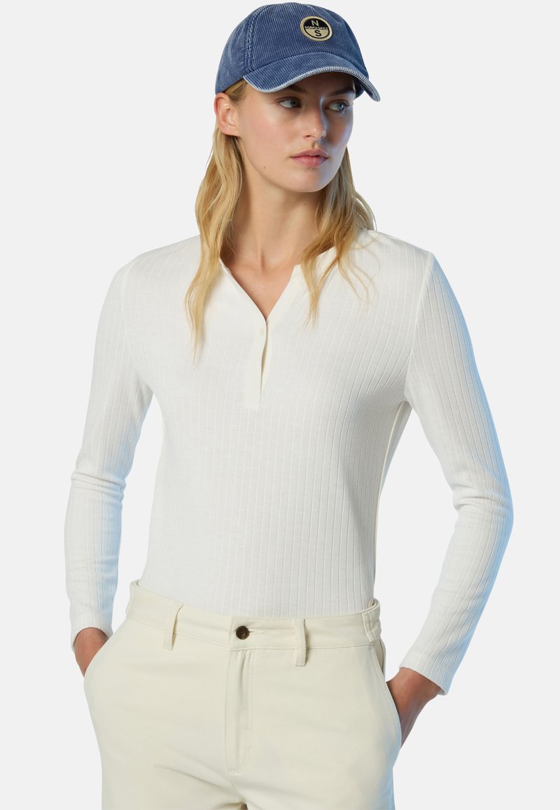 Maglietta bianca a maniche lunghe con collo henley, abbinata a pantaloni beige chiaro. Un berretto blu con logo completa l'outfit.