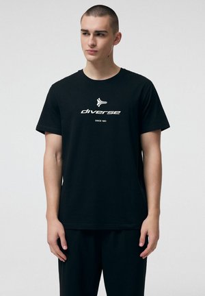 Młody mężczyzna z krótkimi włosami, ubrany w czarną koszulkę z logo i napisem "diverse", stojący na tle jednolitego, jasnoszarego tła.