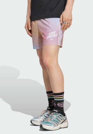 Persona que lleva pantalones cortos con degradado pastel y texto "LOVE UNITES", calcetines negros a rayas y zapatillas Adidas multicolor, de pie frente a un fondo liso.
